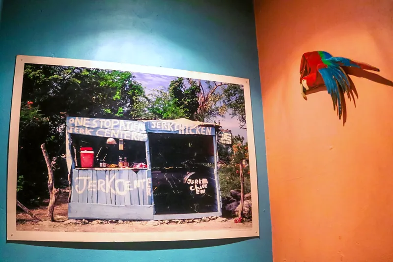 A framed photo of a Jamaican jerk-chicken stall on an orange and aqua wall, with a carved parrot