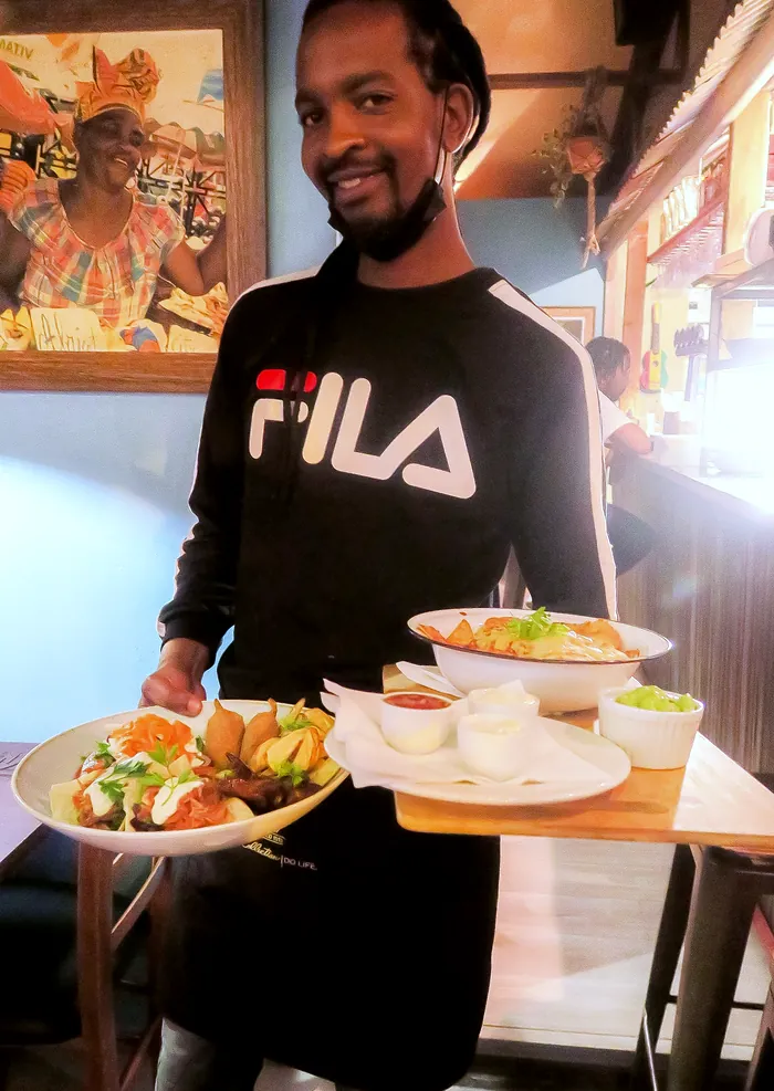 A smiling waiter carrying plates of Caribbean food past a framed portrait
